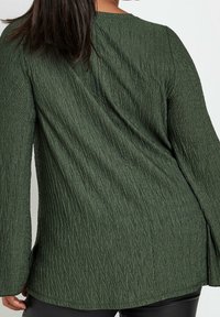 Top verde con texture e un motivo arricciato, con maniche a campana. Il design presenta un collo rotondo e una vestibilità slim in vita.