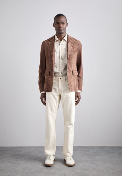 Blazer en daim marron sur une chemise crème à motifs floraux, associé à un pantalon crème et des baskets blanches, se tenant devant un fond neutre.