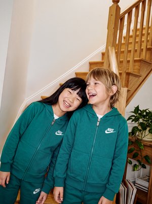 Deux enfants souriants en survêtements Nike assortis de couleur sarcelle se penchent l’un vers l’autre devant un escalier en bois.