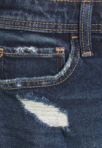 Farkkuhousut tummansinisellä sävyllä, joissa on kulutettu tasku ja valkoinen paikka, korostettuna kontrastivärisellä oranssilla ompeleella.