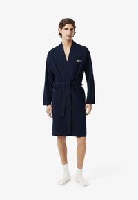 Accappatoio blu navy realizzato in tessuto morbido, con maniche lunghe, una cintura in vita e due tasche frontali. Logo Lacoste sul petto.