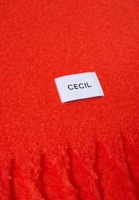 Czerwona chusta z miękkiego materiału, z frędzlami na brzegach. Na powierzchni widoczna jest biała metka z napisem "CECIL".