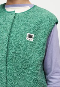 Πράσινο φλις γιλέκο με υφή και κουμπιά που κλείνουν με τρουκς, φορεμένο πάνω από ιώδες μπλουζάκι με μακριά μανίκια, με μαύρο σχέδιο ήλιου στο στήθος.