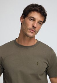 T-shirt en coton vert olive avec un col rond et des manches courtes. Détail notable : petit logo brodé sur la poitrine.