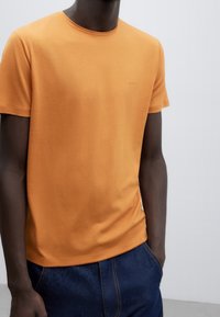 Oranžové bavlněné tričko s kulatým výstřihem, krátkými rukávy a hladkou texturou. Obsahuje jemně vyšitý značkový logo na hrudi.