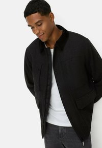 Schwarze Wollmischungjacke mit Kragen, Fronttaschen und Reißverschluss. Verfügt über ein strukturiertes Gewebe und eine lockere Passform. Wird über einem weißen Hemd getragen.