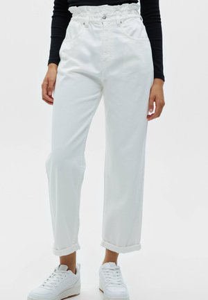 Frau trägt weiße, hochtaillierte Hose mit umgeschlagenem Saum und weiße Turnschuhe, dazu ein schwarzes Langarmoberteil, steht vor einfarbigem Hintergrund.