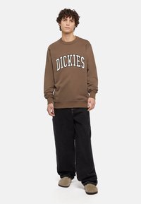 Felpa marrone con la scritta "DICKIES" in lettere bianche, jeans neri larghi e scarpe beige slip-on. Capelli ricci e sfondo neutro.