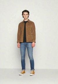 Braune Cordjacke mit Knopfleiste, einer Brusttasche, schwarzes T-Shirt, blaue Jeans und beige Sneakers mit weißen Sohlen.