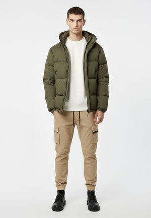 Pikowana kurtka puchowa w oliwkowej zieleni z kapturem, beżowe spodnie cargo z bocznymi kieszeniami i czarnymi akcentami, czarne buty.