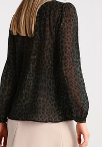 Blouse noire transparente à imprimé léopard vert, manches bouffantes et poignets élastiques. Tissu léger avec une superposition texturée.