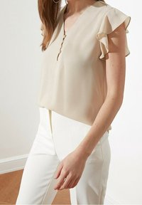 Beige Bluse aus leichtem Stoff, mit V-Ausschnitt und gerafften kurzen Ärmeln. Verfügt über ein Knopfdetail am Ausschnitt. Getragen mit weißen Hosen.