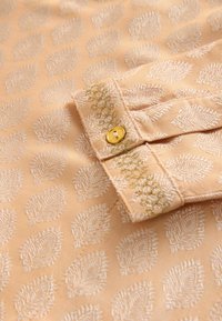 Beige tkanina s potiskem listů a vyšívanými akcenty. Obsahuje zlatý knoflík a dekorativní stehy blízko manžety. Hladká textura.