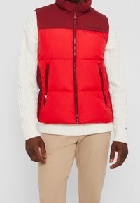 Hombre que lleva un chaleco sin mangas acolchado rojo sobre un suéter de punto blanco y pantalones beiges, de pie contra un fondo claro.