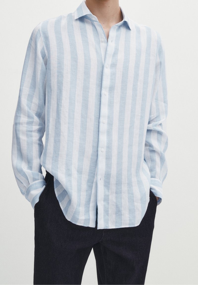 Personne portant une chemise à rayures verticales bleu clair et blanche à manches longues et un pantalon foncé, les mains dans les poches, haut du corps visible.