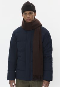 Tumedast sinisest puffer-jakist, kudumist pähe sobivast pruunist beanie'ist ja pika pruuni frangebändiga sallist. Modell poseerib ühtlase valge tausta vastu.