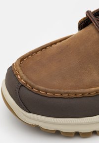 Chaussure décontractée en cuir marron avec une tige texturée, une semelle en caoutchouc gris foncé contrastante et des coutures visibles le long des bords.
