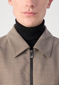 Karirana jakna na zip s smešnim in črnim vzorcem, ki ima zložen ovratnik in črn pulover z visokim ovratnikom spodaj. Vidna je zadrga.