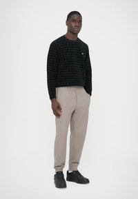 Schwarzer Pullover mit geometrischem Muster, kombiniert mit hellbeigen, strukturierten Hosen. Modell trägt schwarze Turnschuhe.