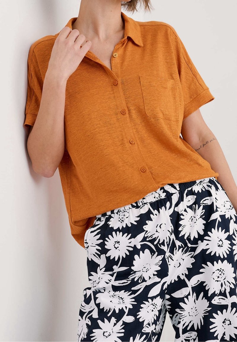 Personne portant une chemise orange ample à boutons et un pantalon noir avec de grands motifs floraux blancs, main près du col, tatouage visible sur le bras.