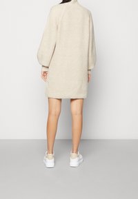 Robe pull en maille beige avec un col montant, des manches oversize et une coupe décontractée, associée à des baskets blanches avec une touche beige.