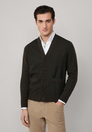 Junger Mann, der einen dunkelgrünen, geknöpften Cardigan über einem weißen Hemd und beigen Hosen trägt, steht mit einer Hand in der Tasche vor einem schlichten Hintergrund.