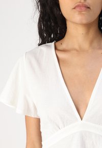 Hvit bomullstop med dyp V-hals, korte volangermammer og en tettsittende bodice med horisontale sømdetaljer.