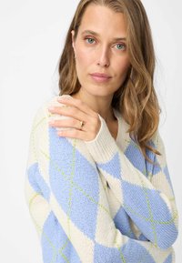 Maglione lavorato a maglia color crema con motivo a rombi blu e verde chiaro, con scollo a V ampio e polsini a coste, indossato da una persona.