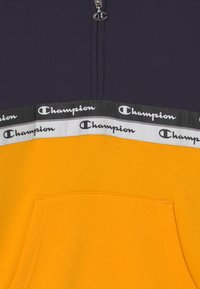Sudadera de medio cierre en algodón de mezcla azul marino y amarillo. Presenta un bolsillo frontal y una banda decorativa con el logo de "Champion" en colores contrastantes.