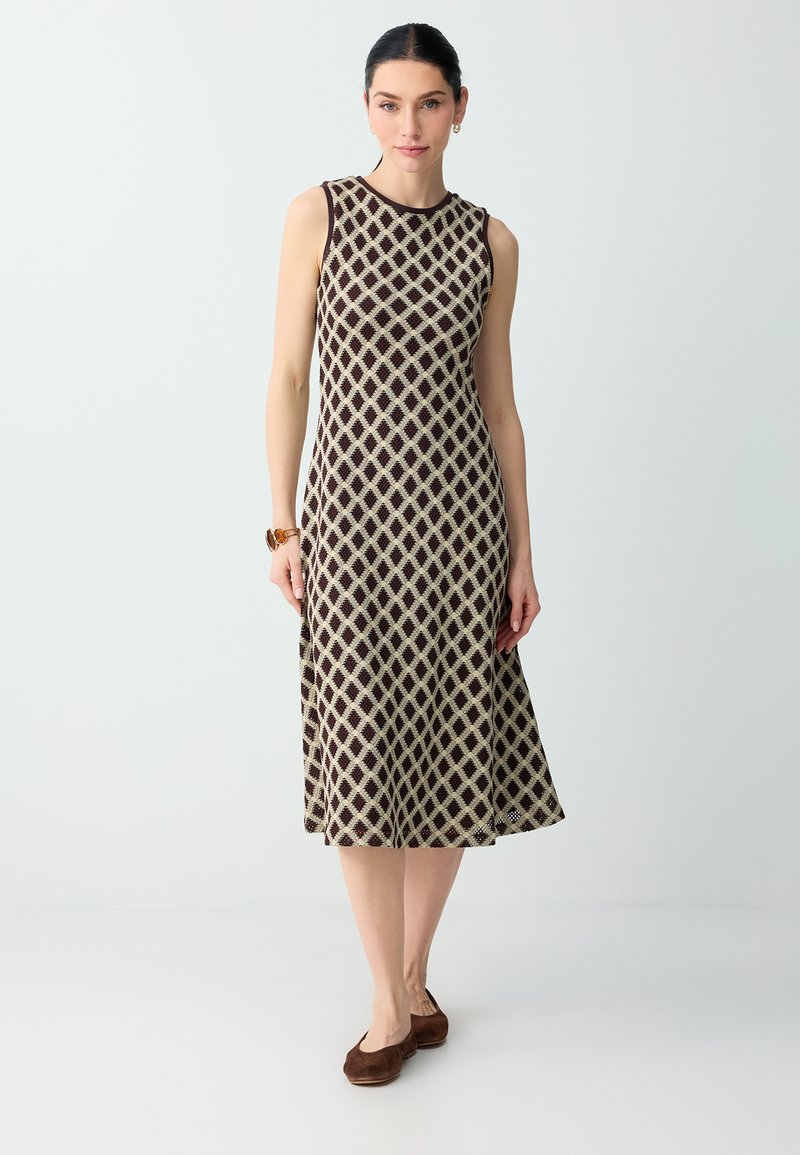 Femme debout portant une robe midi sans manches marron et beige à motif losanges, des ballerines marron, un bracelet et des boucles d’oreilles créoles.