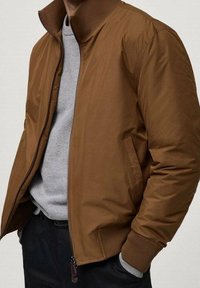 Veste aviateur marron avec col montant, fermeture  éclair et poignets côtelés. Portée sur un sweat-shirt gris clair, avec une texture lisse.