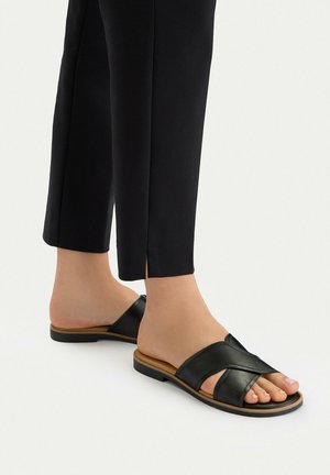 Osoba ubrana w czarne, krótkie spodnie i czarne skórzane, płaskie sandały z krzyżującymi się paskami na jasnym tle.