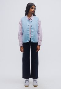 Ανοιχτό μπλε φλις γιλέκο με στρογγυλή λαιμόκοψη και κουμπιά στο μπροστινό μέρος, φορεμένο πάνω από ένα ριγέ ροζ πουκάμισο. Συνδυασμένο με σκούρα τζιν φαρδιάς γραμμής και γκρι παπούτσια.