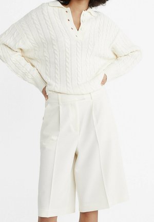 Personne portant un pull torsadé écru avec des boutons et un short à taille haute, jambes larges et longueur genou, posant avec les mains sur les hanches.