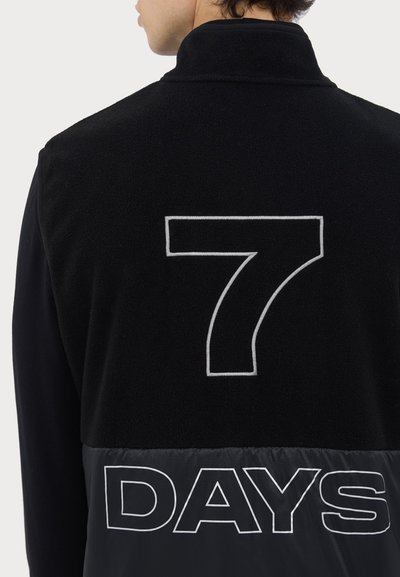 Veste en polaire noire avec un col haut, un grand "7" brodé en blanc à l'arrière et un bas en nylon noir contrastant avec le texte "DAYS".