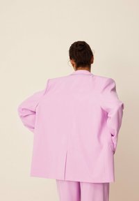 Różowa, oversized marynarka o zdefiniowanej formie, z szerokimi rękawami i pojedynczym rozcięciem z tyłu. Gładki materiał i dopasowane detale.