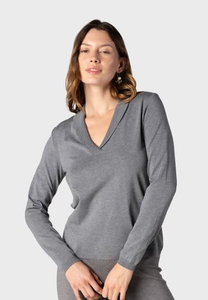 Femme aux cheveux châtain clair portant un pull gris à manches longues en V et un pantalon assorti, debout devant un fond clair uni.