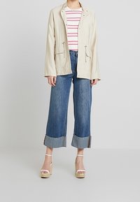 Giacca leggera beige con tasche frontali, abbinata a una camicia a righe bianca e rossa. Pantaloni cropped in denim blu larghi con orlo sfilacciato.