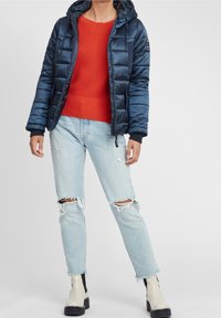 Chaqueta de abrigo azul marino con capucha, con detalles acolchados. Llevada sobre un suéter de punto rojo, combinada con jeans desgastados de un azul claro y botas beige.