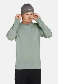 Langärmliges, mintgrünes Shirt mit einer Tasche und einem Rundhalsausschnitt, kombiniert mit einer grauen Strickmütze mit Logo. Weicher Stoff und lockere Passform.