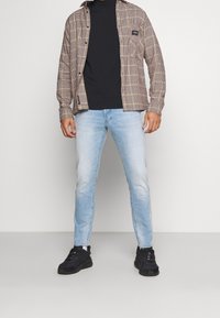 Braun kariertes Hemd, schwarzer Rollkragenpullover, hellblaue Jeans und schwarze Sneaker. Das Hemd hat eine Brusttasche und eine Knopfleiste.