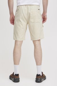 Shorts beiges arrivant au genou avec une taille élastique et une poche arrière. Le tissu semble léger et texturé, associé à des chaussures de sport noires.