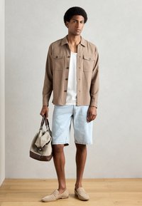 Cămașă bej cu nasturi și două buzunare pe piept, tricou alb dedesubt, pantaloni scurți din denim bleu deschis, mocasini bej și o geantă tote maro cu bej.