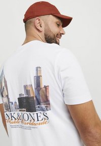 Weißes T-Shirt mit einem großen Stadtbild-Print auf der Rückseite, kurzen Ärmeln und einem klassischen Rundhalsausschnitt. Ergänzt durch eine rostfarbene Mütze.