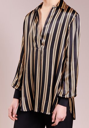 Vrouw die een zwart-beige vertical gestreepte blouse met lange mouwen draagt, met een diepe V-hals en zwarte manchetten.