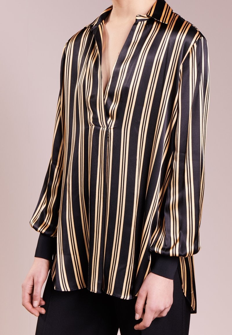 Femme portant un chemisier à manches longues rayé verticalement noir et beige avec un col en V profond et des poignets noirs.