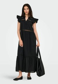 Black maxi dress features ruffled sleeves, lace accents, and a tiered hem. Paired with black flats and a large tote bag. Smooth cotton fabric.