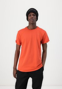 Oransje bomull T-skjorte med rullede ermer, rund hals og liten logo detaljer. Kombinert med svarte bukser, står modellen mot en ensfarget bakgrunn.