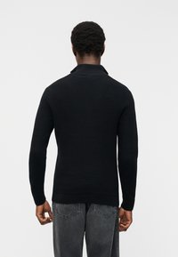 Άνδρας που στέκεται με την πλάτη προς την κάμερα, φορώντας μαύρο μακρυμάνικο πουλόβερ με υφή και ξεθωριασμένο μαύρο τζιν, πάνω σε απλό φόντο.