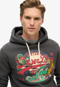 Sudadera con capucha de color gris oscuro que presenta una impresión gráfica de un dragón y un tigre, con colores contrastantes y capucha con cordones. Tejido suave y texturizado.